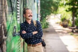 Hamilton Nickoloff, a young man with his haired pulled back leans against a colorful wall mural, smiling